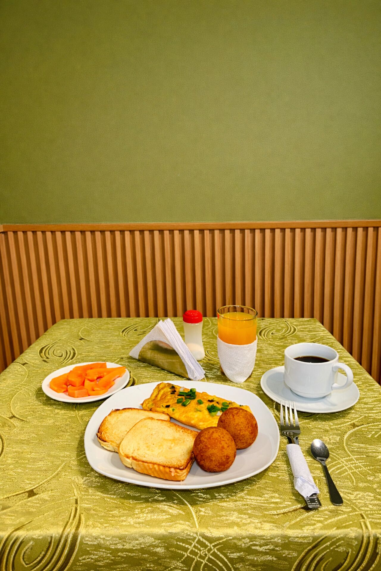 Una mesa de comedor con mantel verde, un plato de comida que incluye tostadas, una tortilla, albóndigas, un vaso de jugo de naranja, una taza de café y una botella de leche.