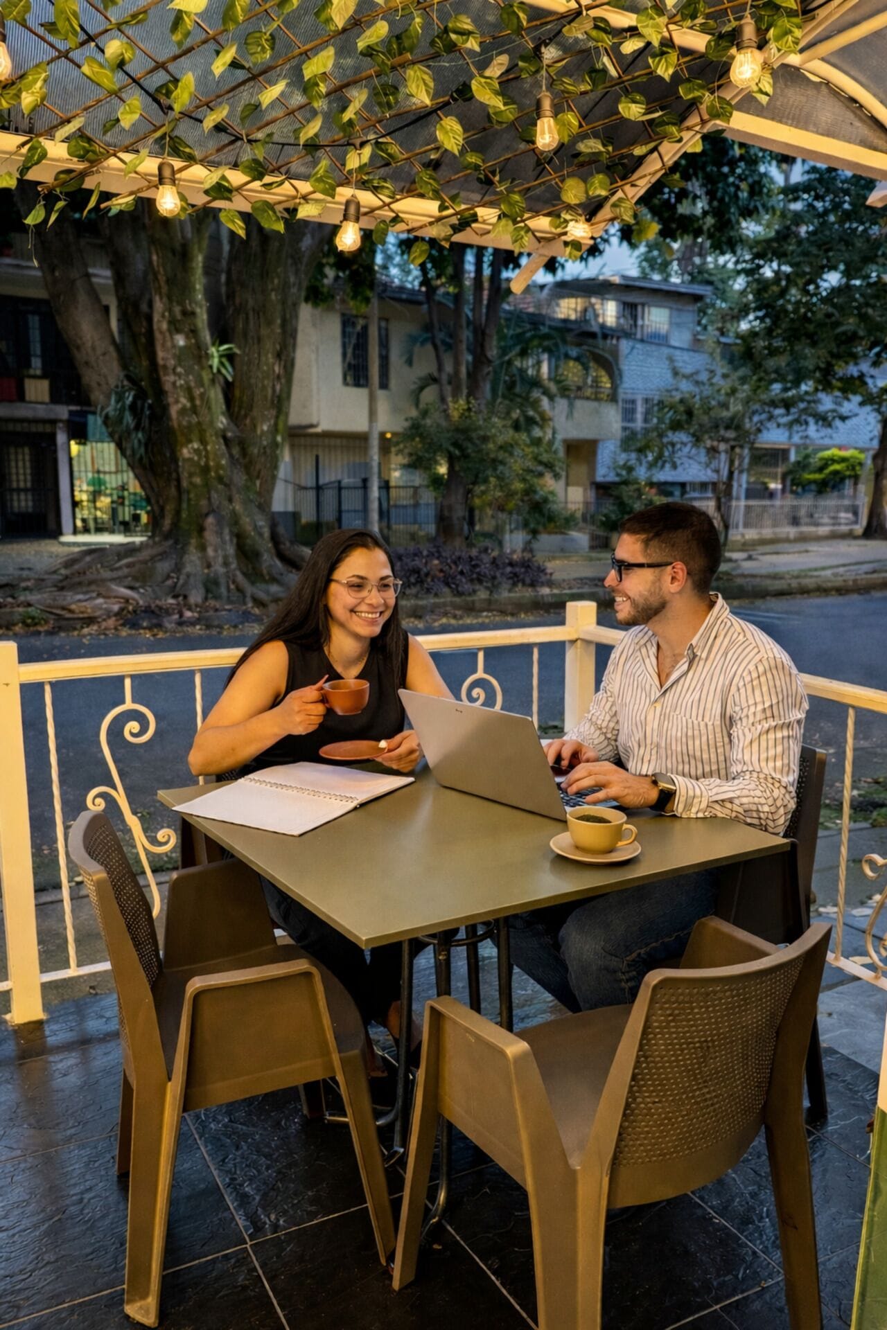 Two people seated at an outdoor table with a laptop, surrounded by chairs and a railing.