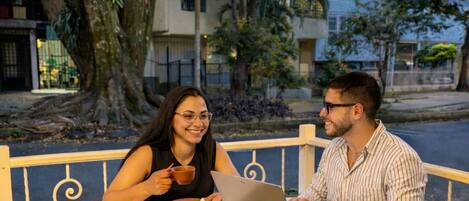 Two people seated at an outdoor table with a laptop, surrounded by chairs and a railing.