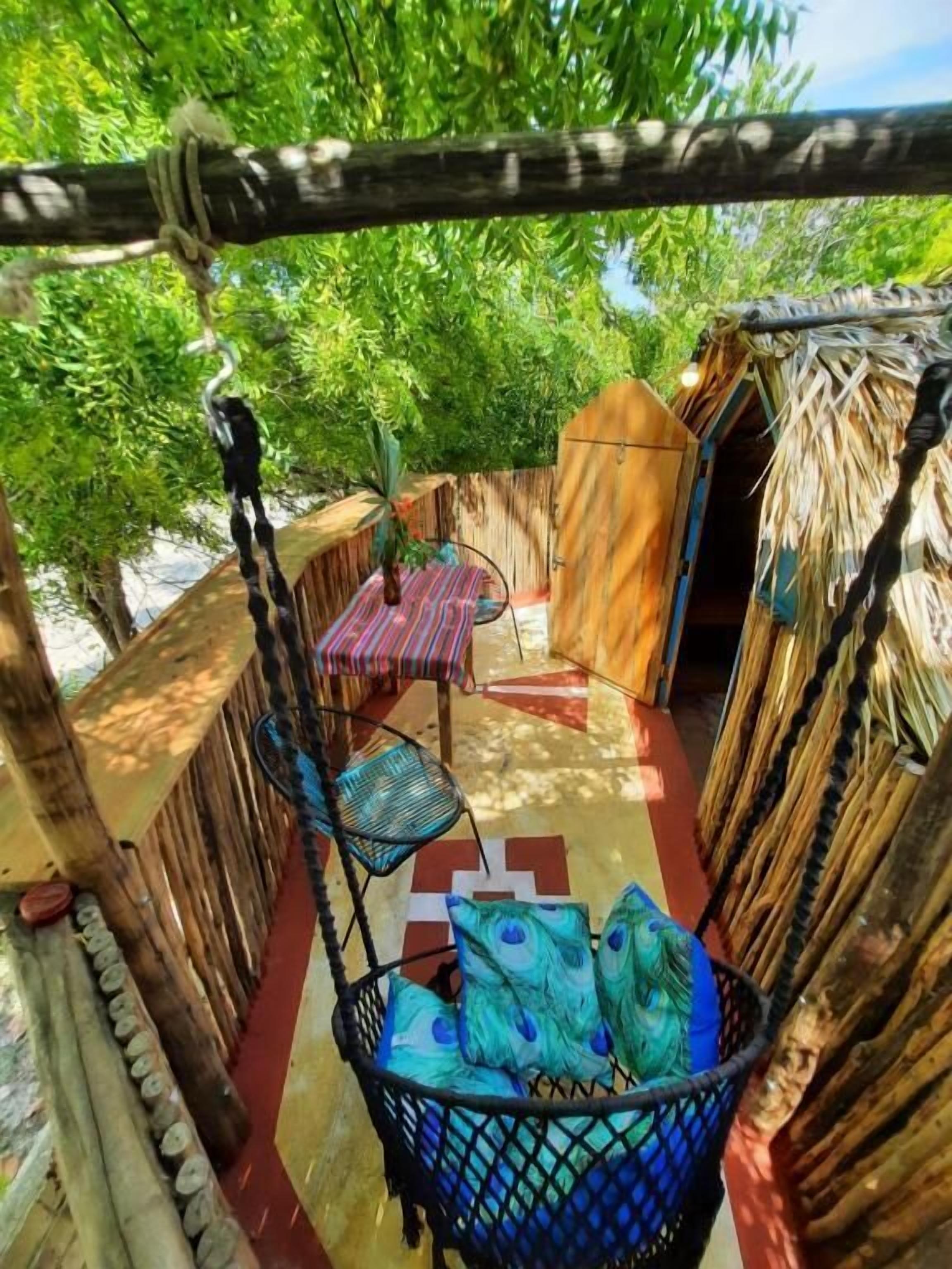 Un espacio de descanso al aire libre con un sillón tejido, una mesita pequeña y una cesta colgante con cojines.