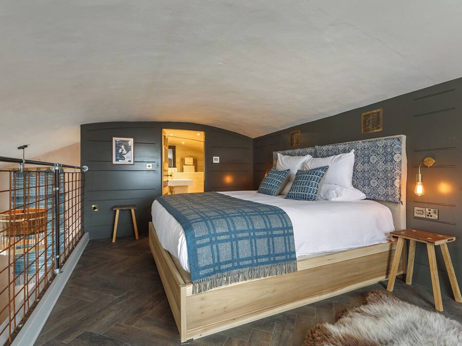 Een slaapkamer met een bed, een houten krukje, een bonttapijt, een hanglamp en een deur.