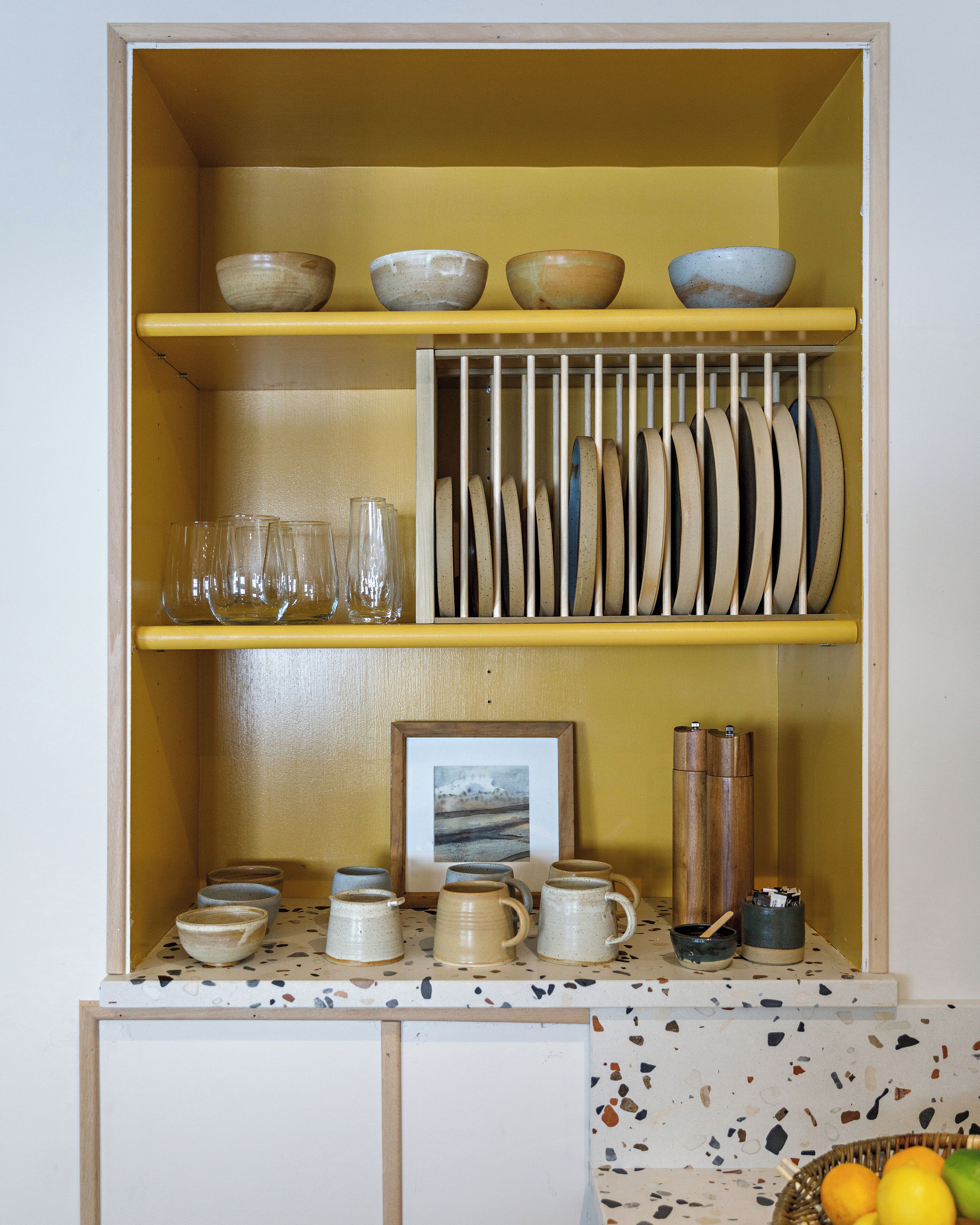 Una cocina con un mueble amarillo que contiene diversos utensilios de cocina, una encimera con un diseño de mosaico y una cesta de frutas.