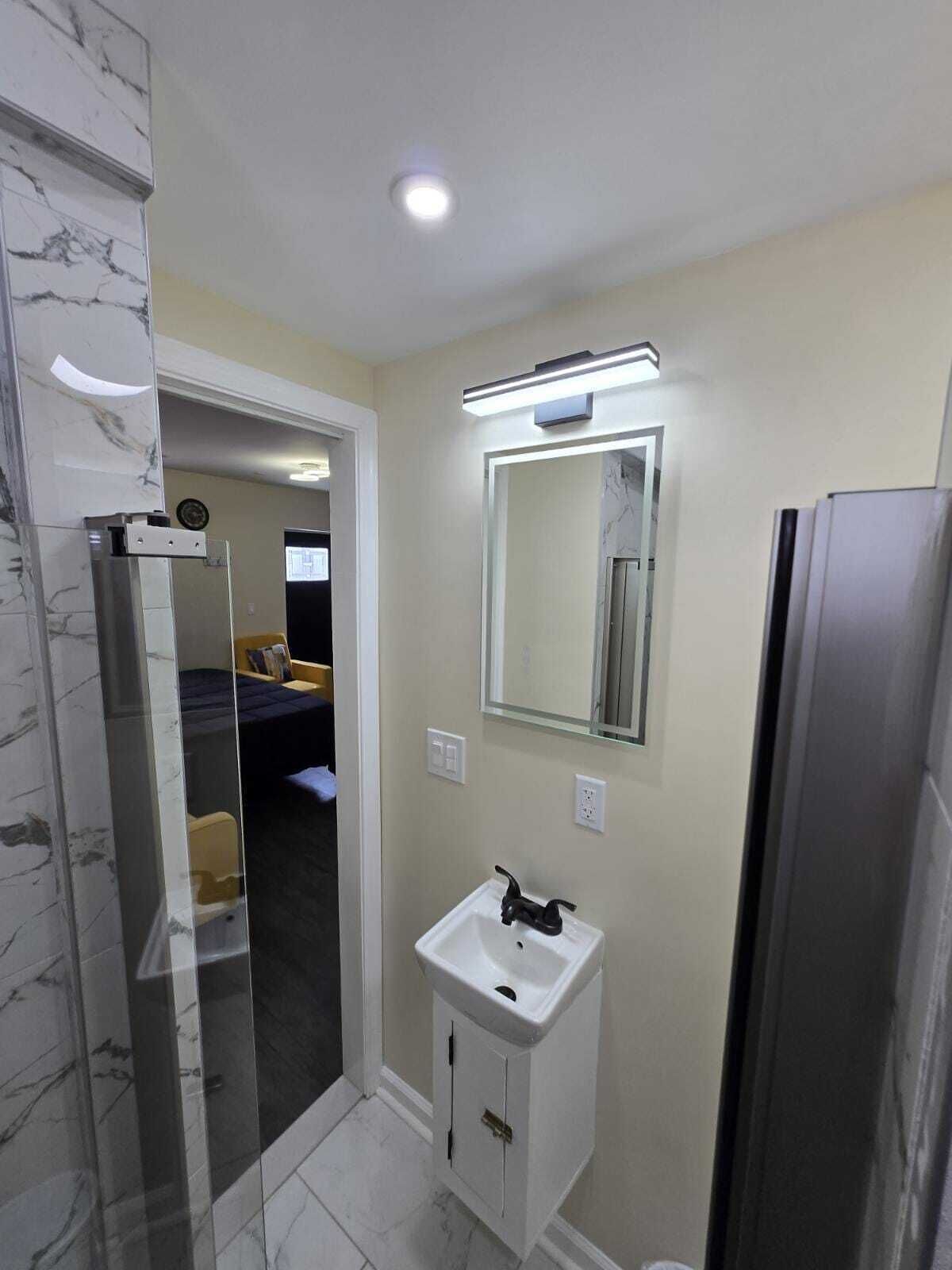 A modern bathroom with a marble wall, a glass shower enclosure, a white sink with a black faucet, and a mirror above it.