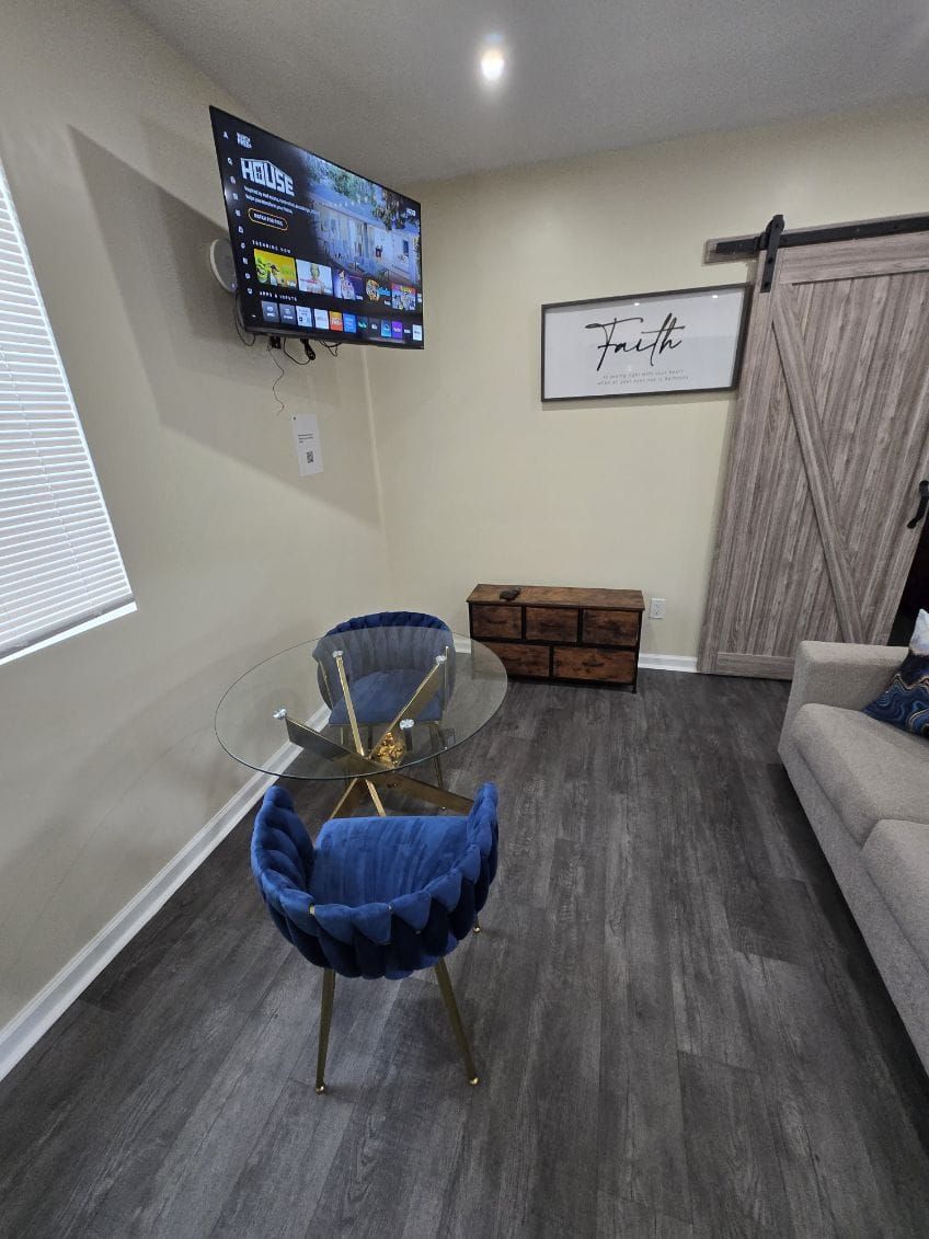 Un salon avec une table en verre, deux fauteuils bleus, un téléviseur fixé au mur et une citation encadrée accrochée au mur.