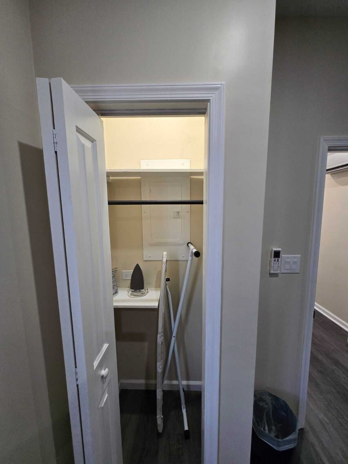 A narrow hallway with a door leading to a small room containing a laundry area with a washing machine, ironing board, and a drying rack.