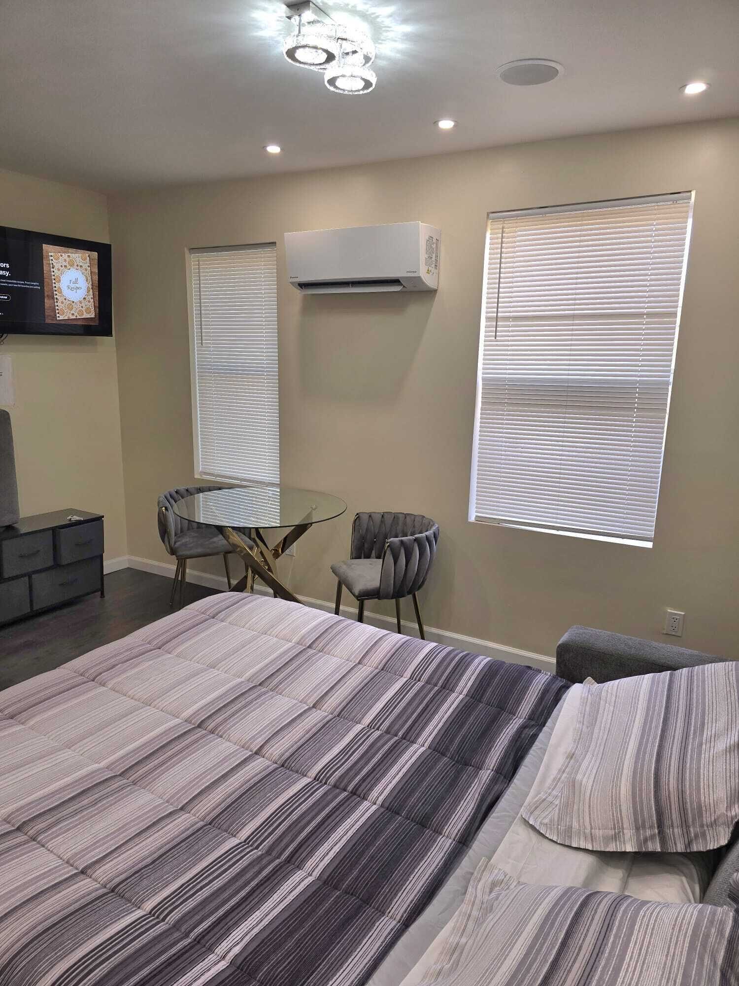 A modern bedroom with a large bed, a desk, two chairs, an air conditioning unit, and a TV on the wall.