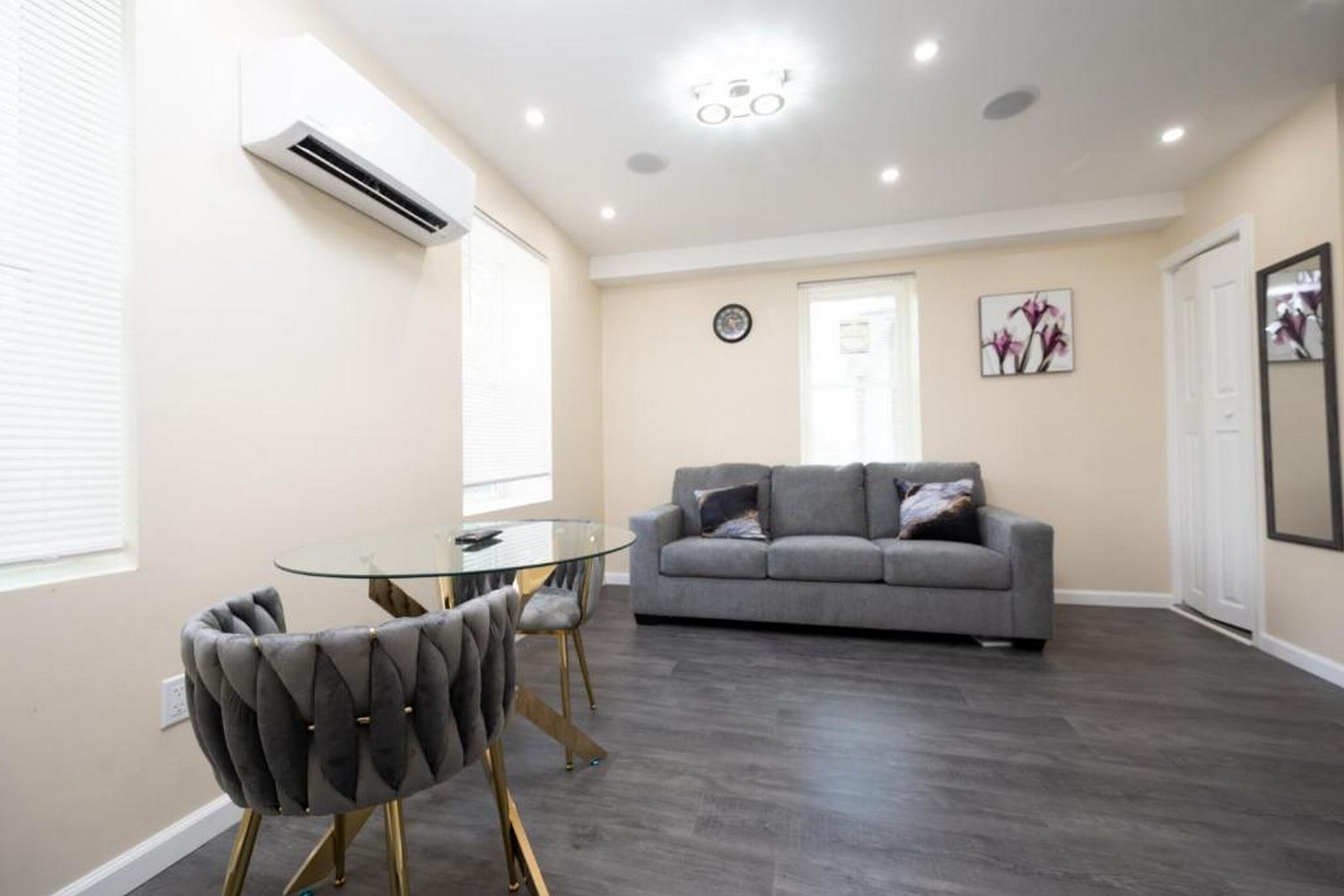 A modern living room with a glass-top dining table, tufted armchairs, a grey sofa, and a wall-mounted air conditioner.