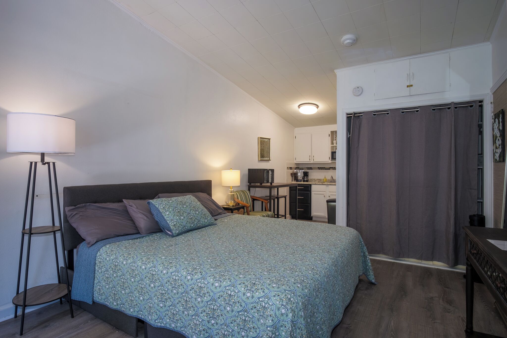 Ein Schlafzimmer mit Bett, Nachttisch, Lampe und einer Küchenzeile mit Schränken und einer Mikrowelle.