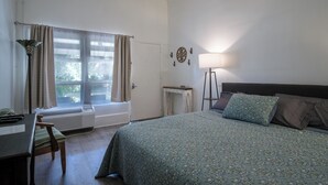 A bedroom with a bed, a desk, a chair, a window with curtains, a radiator, a wall clock, and a floor lamp.