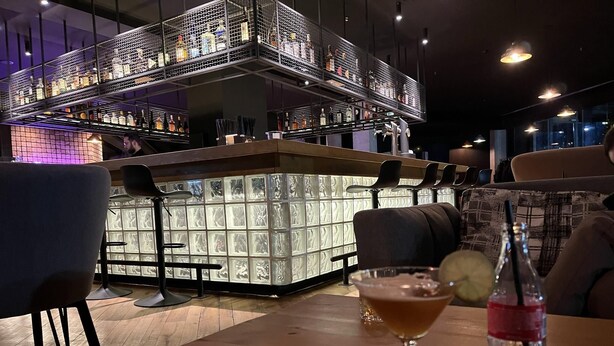 Un espace bar doté d’un comptoir haut, d’étagères remplies de bouteilles et d’un coin salon avec une table et des chaises.
