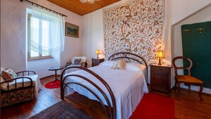 Un dormitorio con techo de madera, una cama con cubrecama blanco, dos mesitas de noche con lámparas, una silla, una pequeña mesa y una ventana con cortina de encaje.