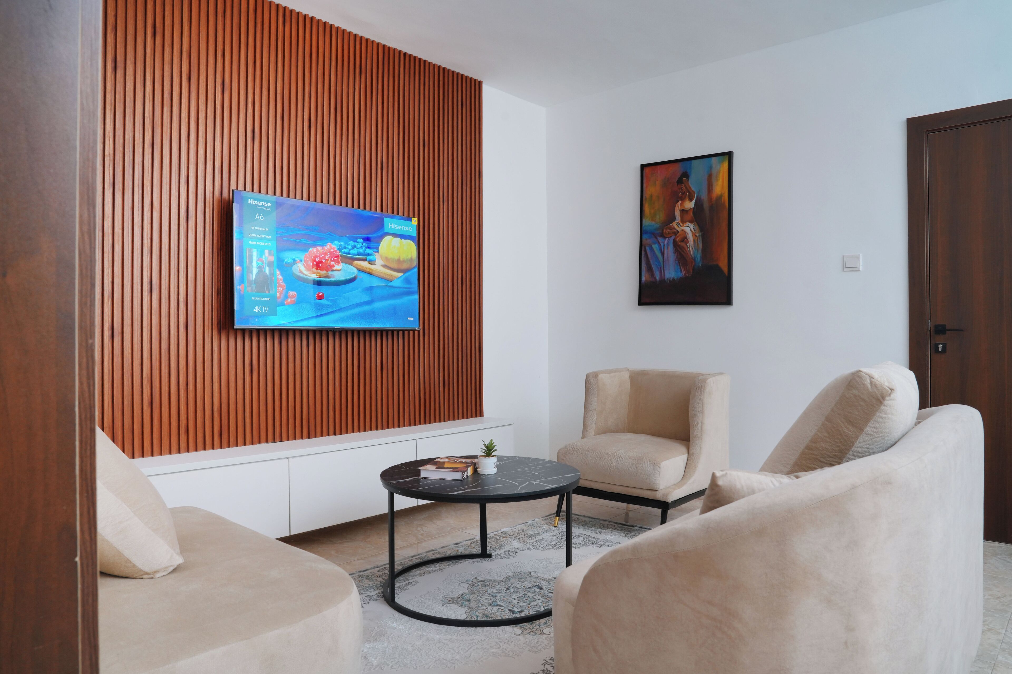 Ein modernes Wohnzimmer mit Flachbildfernseher an einer Wand aus Holzpaneelen, zwei beigefarbenen Sesseln, einem kleinen runden Couchtisch und einem Bild an der Wand.