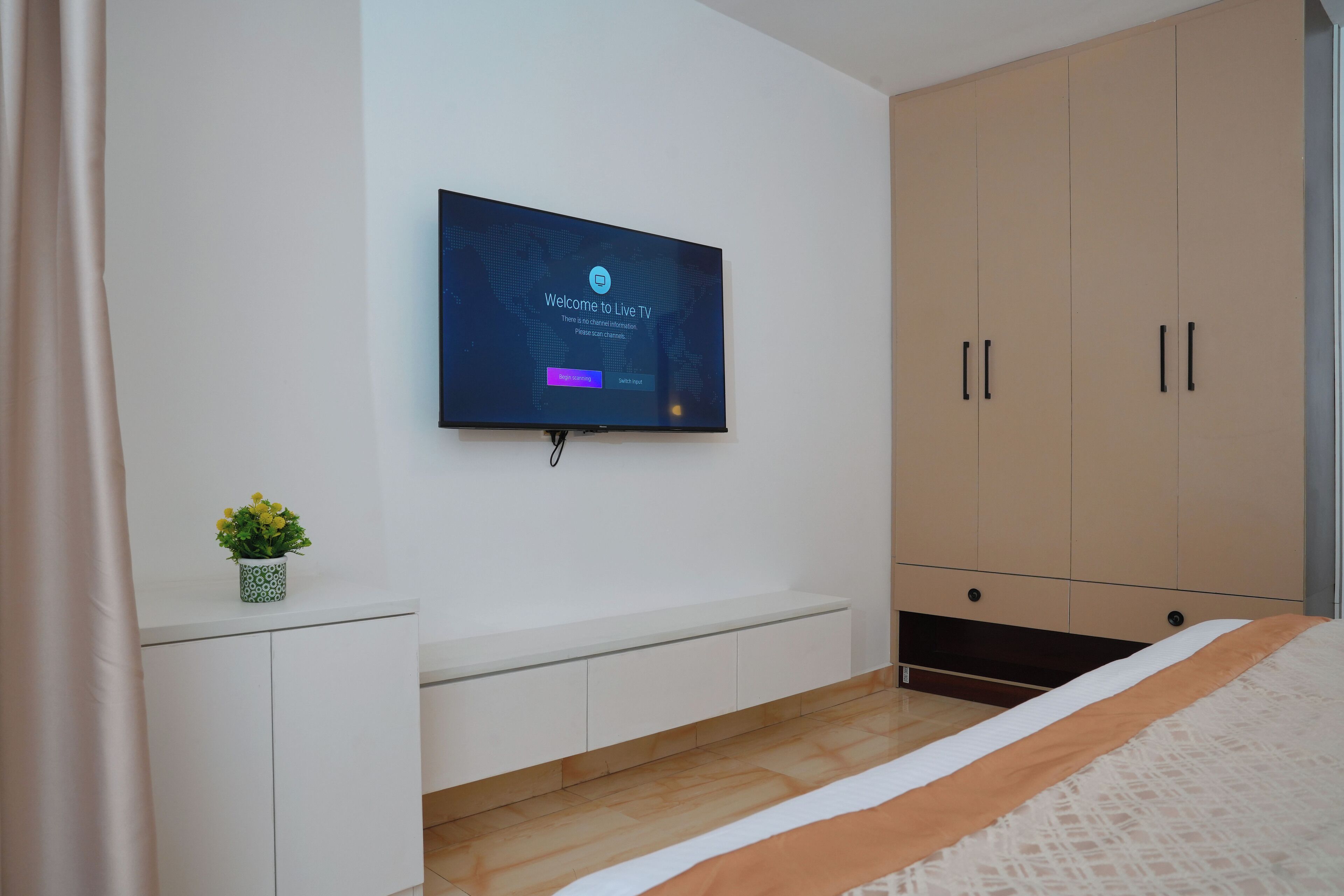 A bedroom with a TV mounted on the wall, a bed with a white and orange bedspread, a small potted plant on a white console table, and a beige wardrobe.