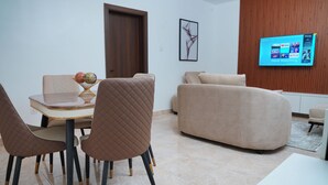 A modern living room with a dining area, a flat-screen TV mounted on the wall, and a wooden panel accent. - Majestic Courts (Lagos)