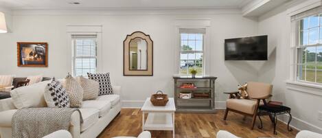 A living room with a white sofa, a glass coffee table, a TV mounted on the wall, and a fireplace with a basket on the mantel.