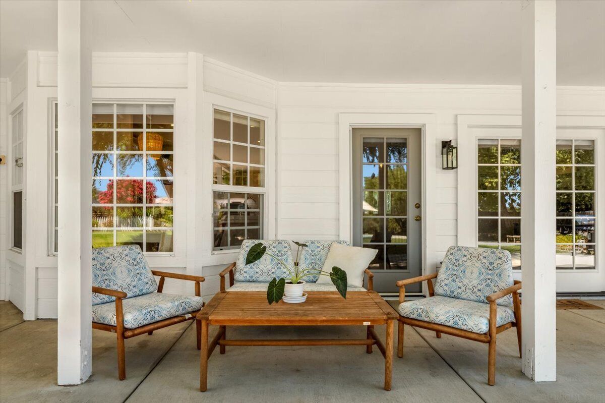 A covered porch with a wooden table and two patterned chairs, large windows, and a door with glass panels.