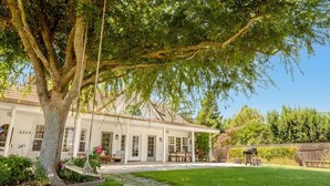 Une maison blanche avec un patio couvert, un chemin de pierre et un grand arbre au premier plan.