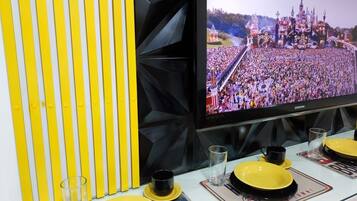 Un televisor muestra una multitud en un evento de castillo, una pared a rayas amarillas y una mesa con platos amarillos y tazas negras.