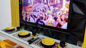 Un televisor mostrando una multitud en un evento, con una mesa delantera donde hay un control remoto, un cuenco y una taza.