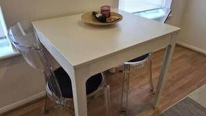 A small, white table with a plate of food and a drink on it, a clear plastic chair, and a window in the background. - Split level luton town center flat (Luton)