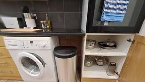 A compact kitchen with a washing machine, a trash bin, and a shelf with pots and pans. - Split level luton town center flat (Luton)