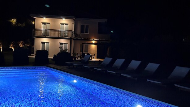 Ein Schwimmbecken mit blauer Beleuchtung, Liegestühlen und einem Gebäude im Hintergrund.