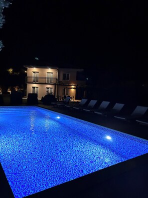 Ein Schwimmbecken mit blauer Beleuchtung, Liegestühlen und einem Gebäude im Hintergrund.