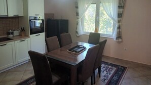 Eine kompakte Küche mit weißen Schränken, einem Esstisch mit sechs Stühlen, einem Kühlschrank und einem Fenster mit transparenten Vorhängen.