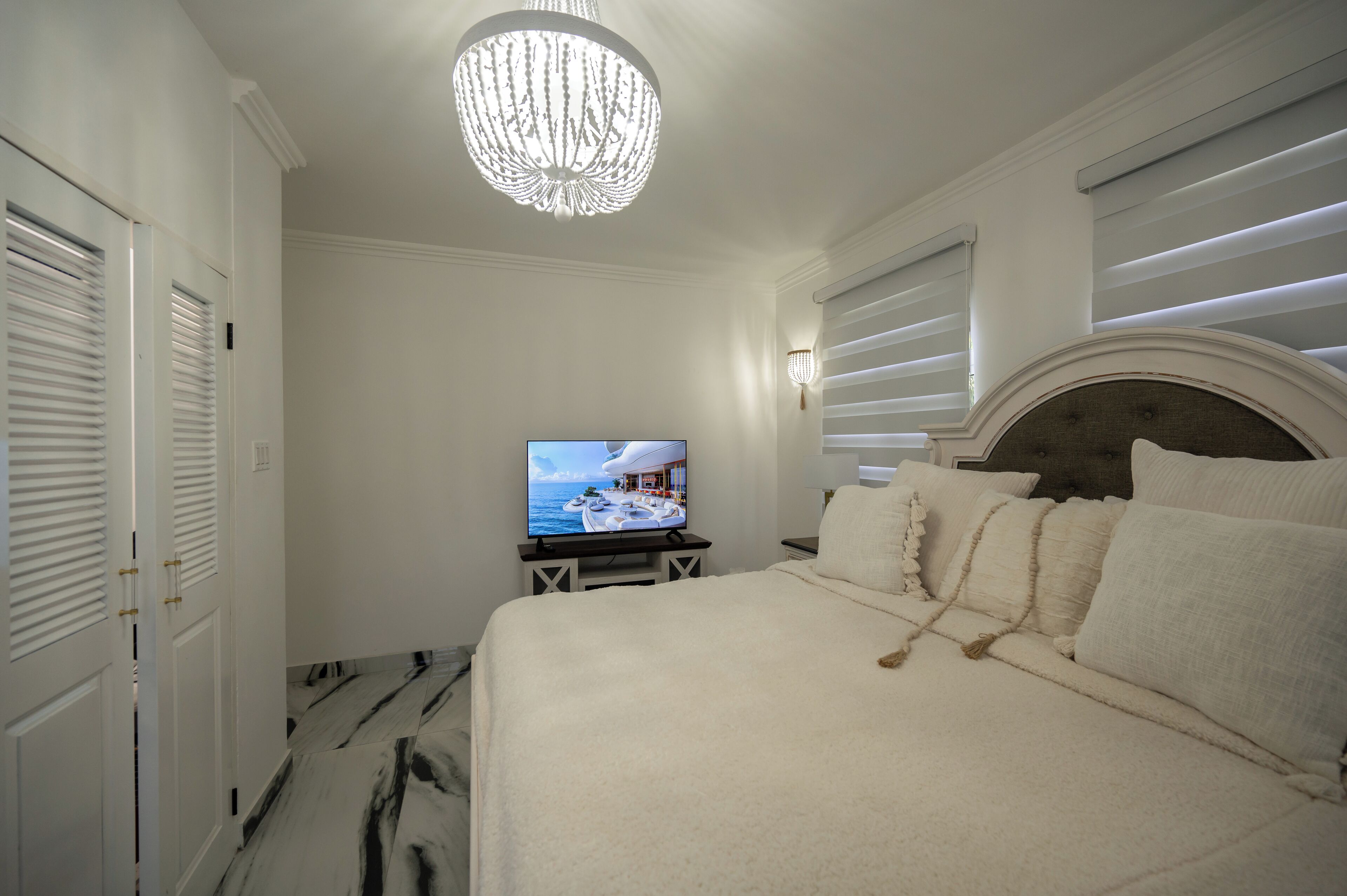 Un dormitorio con cama, televisión, lámpara de araña y ventana con persianas.