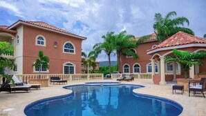 Ein Pool mit Liegestühlen und einer Pergola, umgeben von Palmen und einem Gebäude mit Bogenfenstern und einem Ziegeldach.