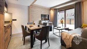 Ein modernes Wohnzimmer mit Sofa, einem Esstisch mit Stühlen, einem an der Wand befestigten Fernseher und einem Balkon mit Vorhängen.