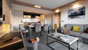 Ein modernes Wohnzimmer mit einem grauen Sofa, einem Couchtisch und einem Essbereich mit Stühlen sowie einer Küche im Hintergrund.
