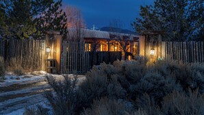 A rustic cabin with a wooden fence, a snow-covered ground, and a warm light illuminating the entrance. - Oasis Escondido / Stunning Views / Spa / Firepit / Location / Private! (El Prado)