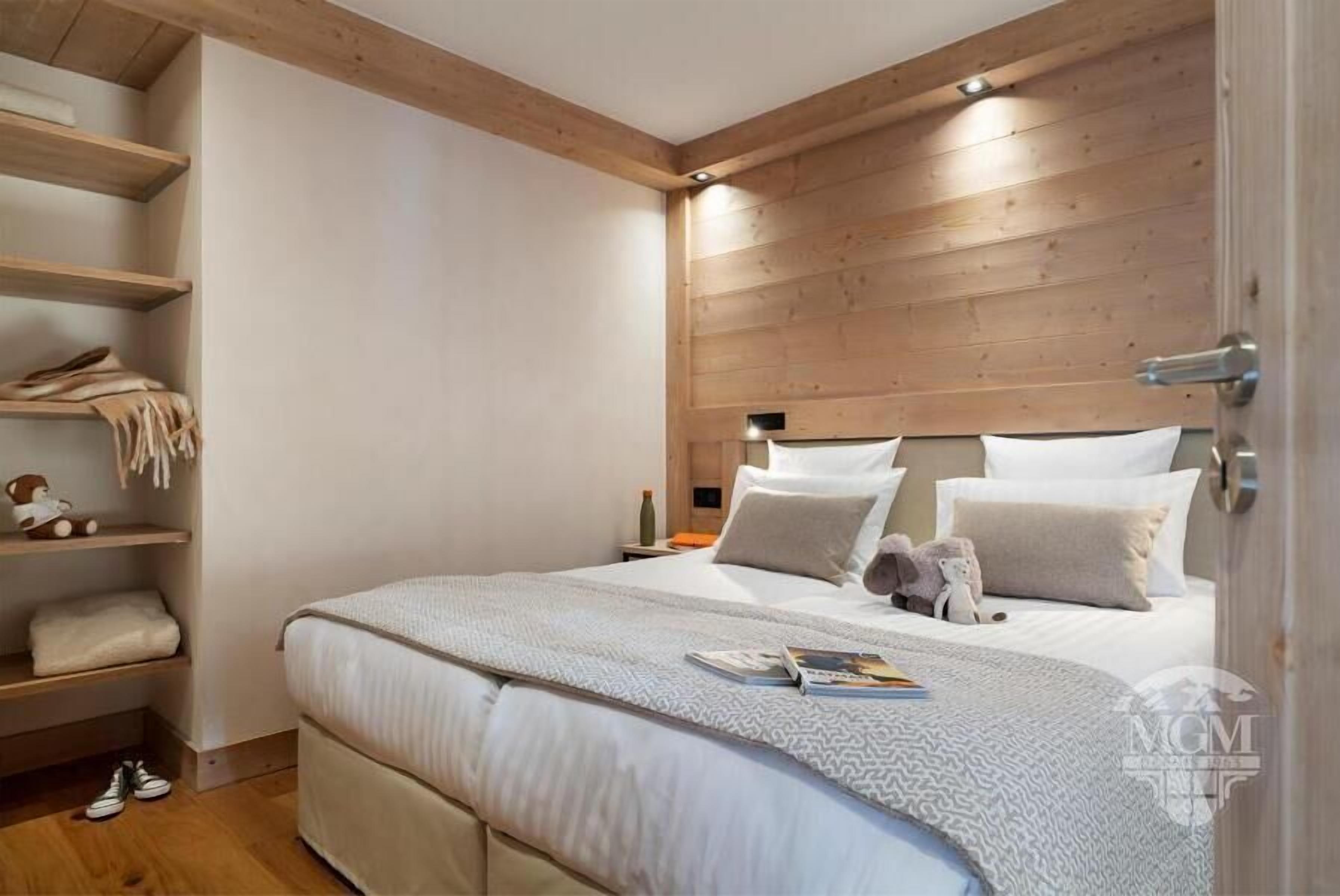 Een slaapkamer met een houten hoofdbord, een bed met witte beddengoed, een nachtkastje met een lamp, en een plank met opgevouwen handdoeken en een teddybeer.