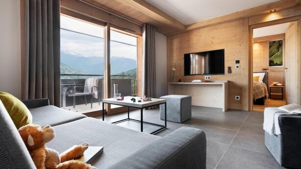 Ein modernes Hotelzimmer mit großem Fenster, einem Flachbildfernseher, einer kleinen Küchenzeile und einer gemütlichen Sitzecke mit einem kuscheligen Stofftier.