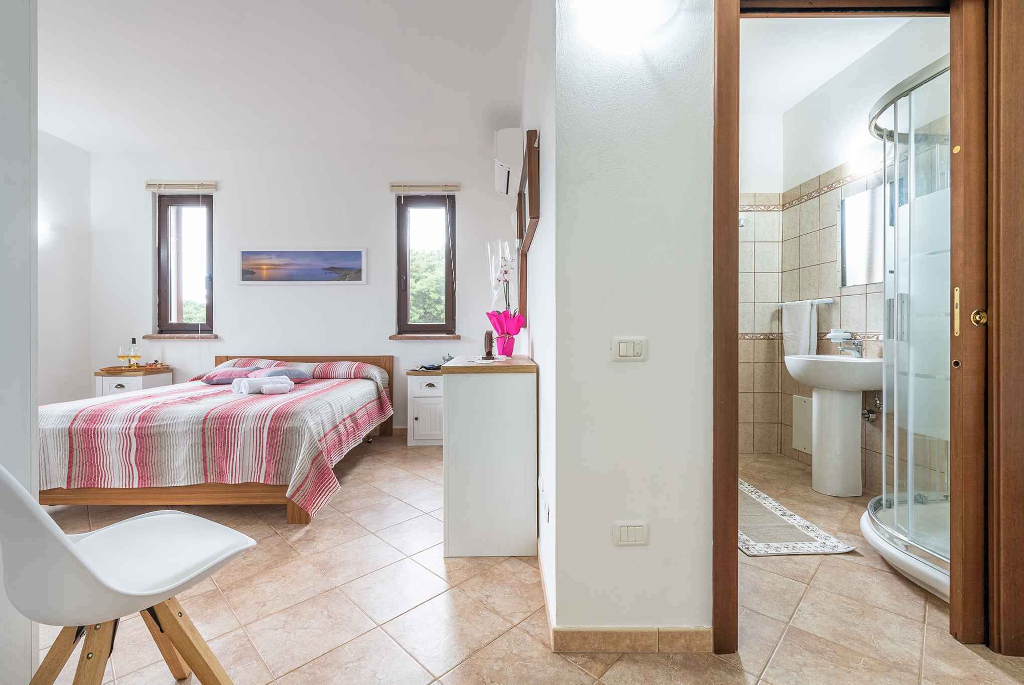 A bedroom with a bed, bedside tables, a chair, and a bathroom with a shower and sink.