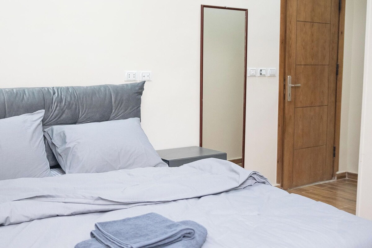 A bedroom with a bed, pillows, a bedside table, a mirror, and a wooden door.
