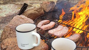 Un feu de camp avec des saucisses en train de griller, deux tasses ornées de citations inspirantes et un paysage rocheux avec des arbres en arrière-plan.
