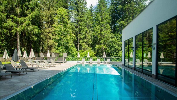 A modern outdoor swimming pool with lounge chairs and umbrellas, surrounded by trees. - Holiday apartment with wellness area (Ötz im Ötztal)