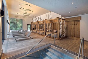 A modern indoor pool area with a wooden floor, lounge chairs, and a pool with steps leading in.