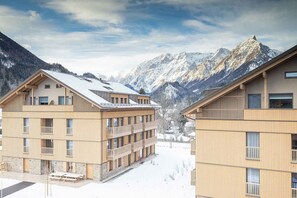 Ein mehrstöckiges Gebäude mit Balkonen, verschneitem Untergrund und Bergen im Hintergrund.