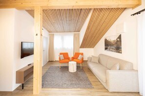 Ein modernes Wohnzimmer mit Fernseher, zwei orangefarbenen Sesseln, einem beigen Sofa und einer Holzbalkendecke.
