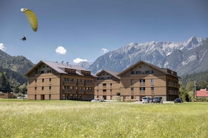 Ein mehrstöckiges Gebäude mit Balkonen, ein Feld im Vordergrund und Berge im Hintergrund.