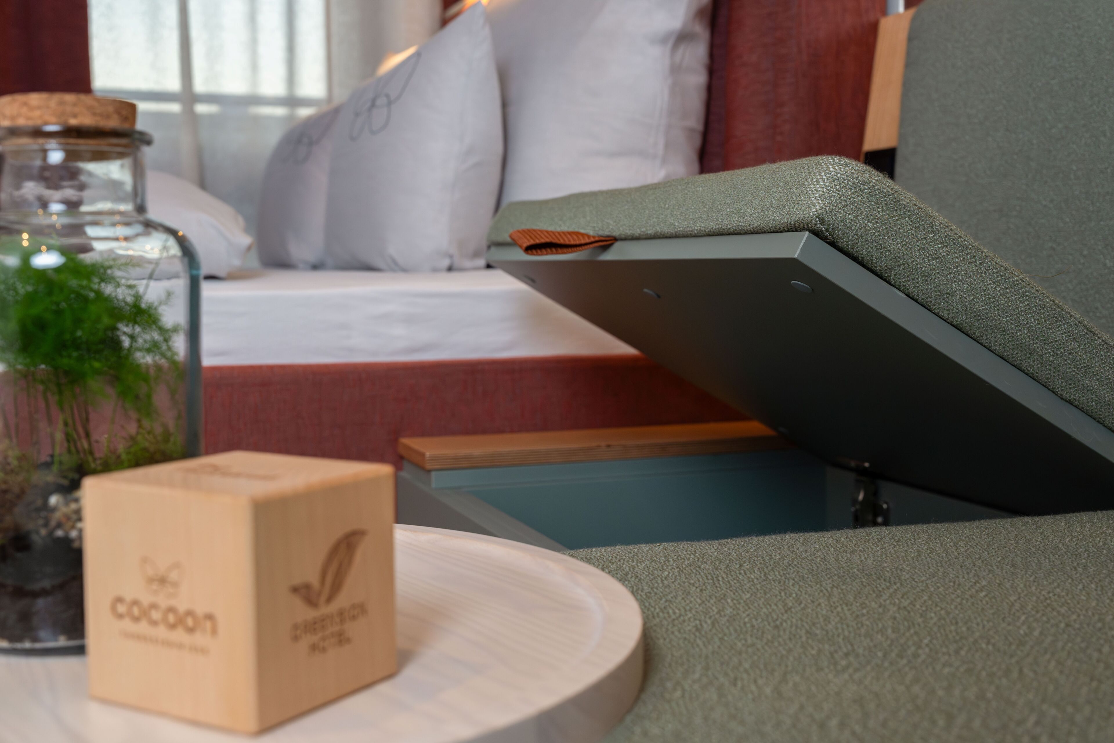 A bed with white pillows, a glass jar with greenery, a wooden box labeled 'cocoon,' and a gray upholstered chair.