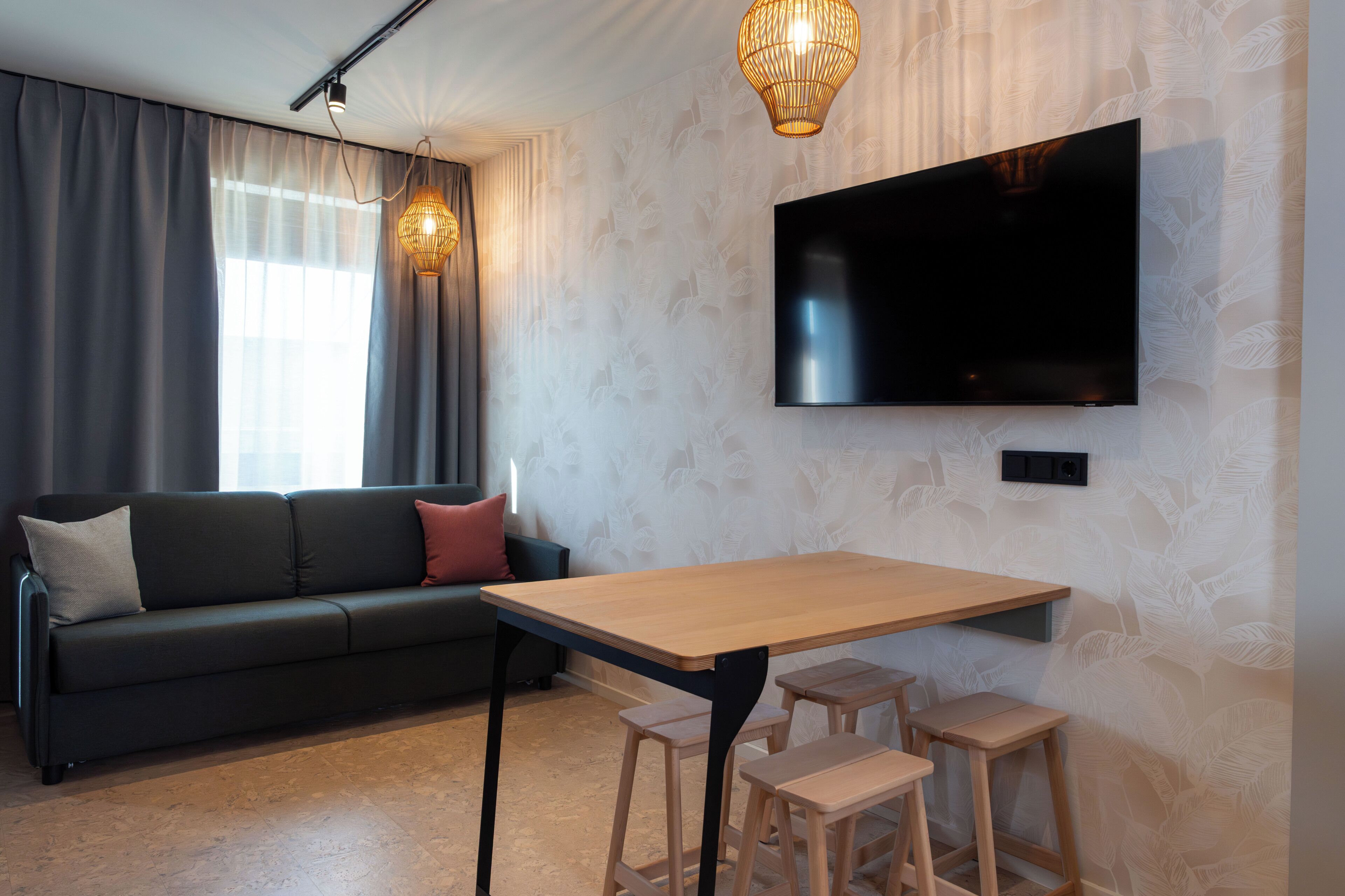 A modern living room with a sofa, a TV mounted on the wall, a wooden table with stools, and a patterned wallpaper.