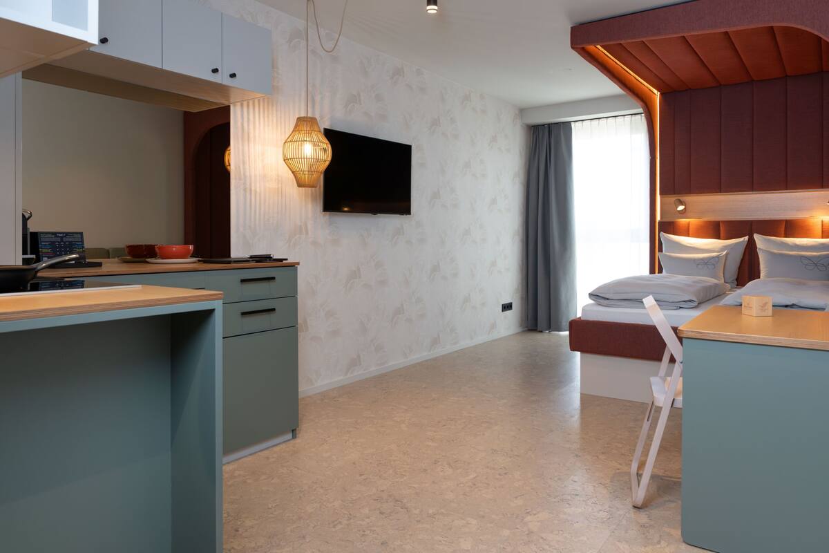 A modern hotel room with a flat-screen TV, a wooden headboard, a bed with pillows, a desk with a computer, and a dining area with a table and chairs.