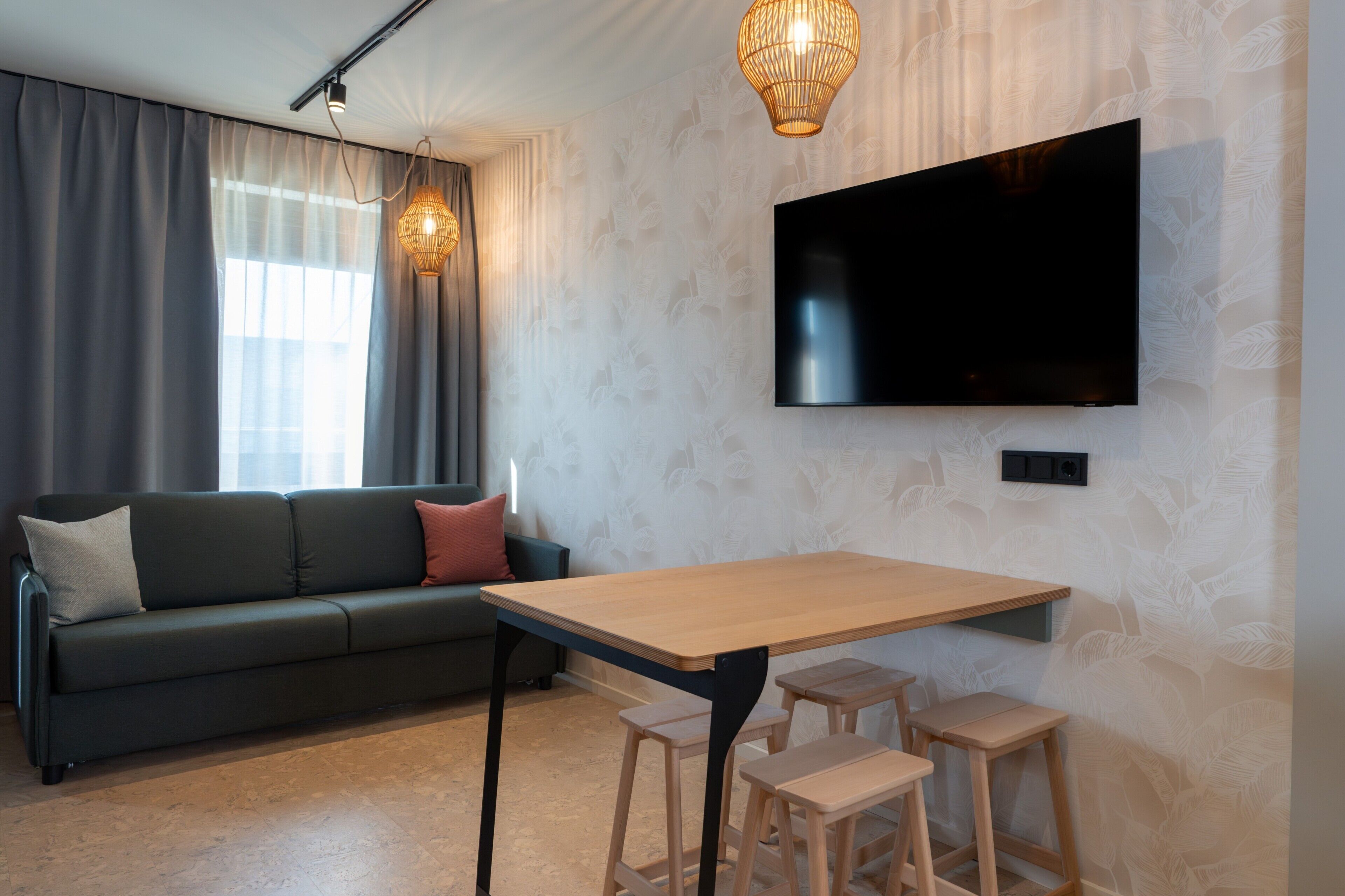 A modern living room with a sofa, a TV mounted on the wall, a wooden table with stools, and a patterned wallpaper.