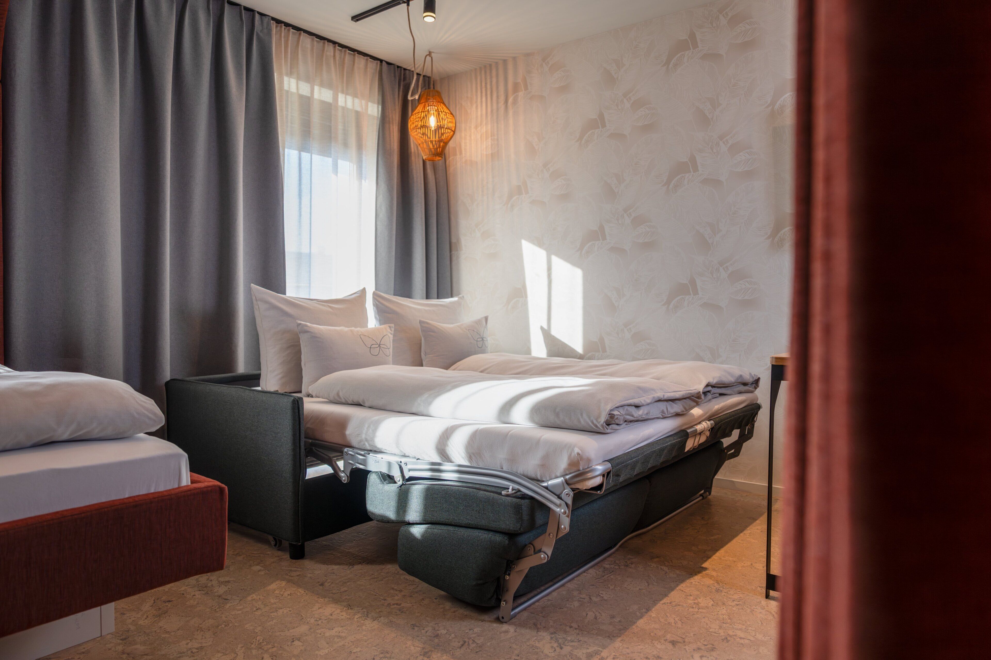Une chambre d’hôtel moderne dotée d’un lit convertible, d’un coin salon séparé et d’une grande fenêtre avec des rideaux.