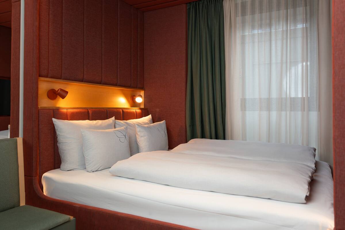 A hotel room with a bed, pillows, a headboard with lights, a green curtain, and a wooden wall panel.