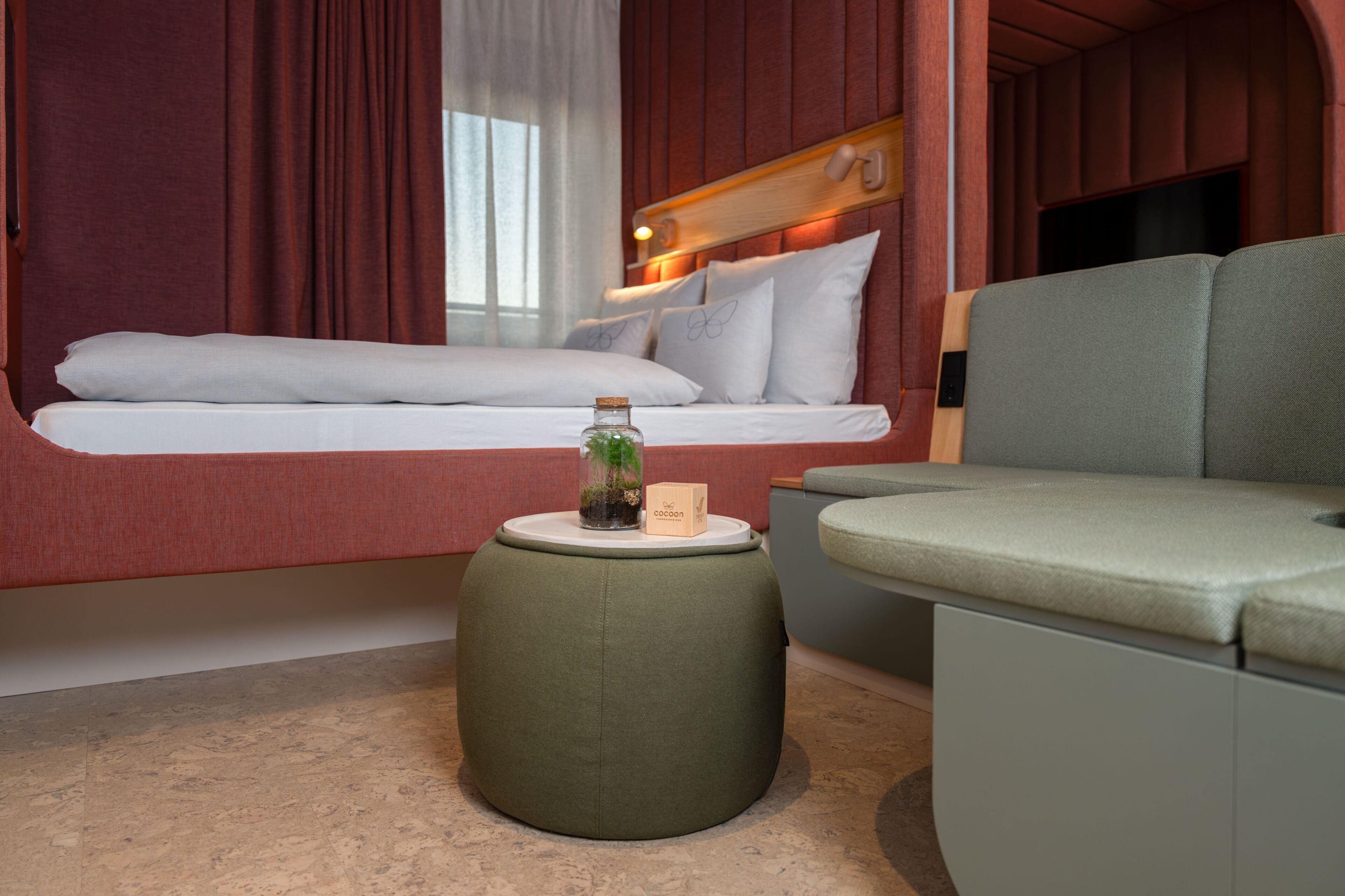 Une pièce comprenant un lit, un pouf vert, une petite table avec une bougie et une plante, ainsi qu’un téléviseur.